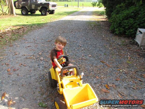 Child sits on a yellow toy tractor with a loader bucket. A camouflage Ford Bronco is parked in the background