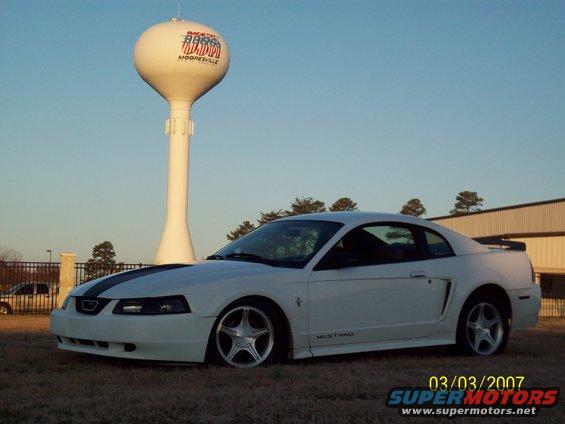 White 2000 Ford Mustang coupe with black racing stripes on hood, parked outdoors near a water tower