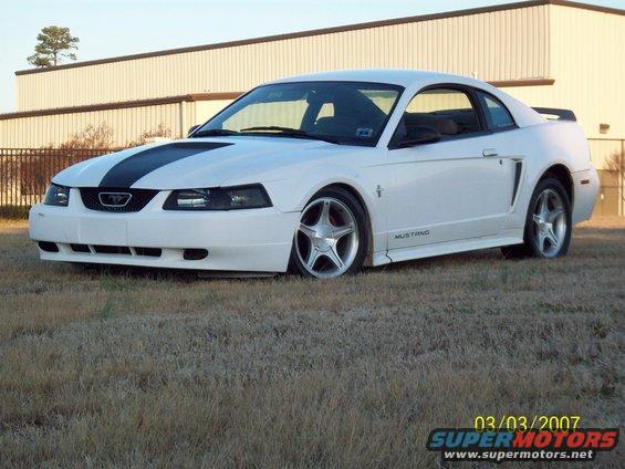 White 2000 Ford Mustang with black racing stripes on hood, parked on grass in front of a building
