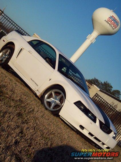 White 2000 Ford Mustang coupe with black racing stripes parked on grass, water tower in background