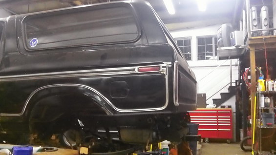 Black Ford Bronco in a garage, rear quarter view. It's on a lift with tools and shelves in the background