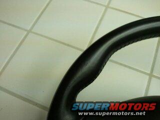 Close-up of a black leather-wrapped steering wheel rim with stitching. Watermark reads SUPERMOTORS.NET