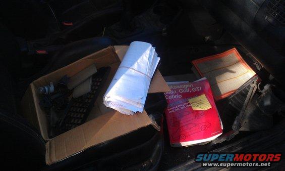 Car interior with a box of parts, stacked papers, an air filter, and a red manual for Volkswagen Jetta, Golf, GTI, and Cabrio