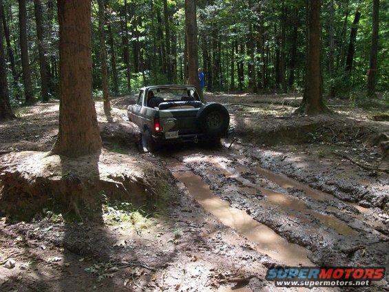 Dark green 1978 Ford Bronco driving through a muddy forest trail, spare tire mounted on the rear