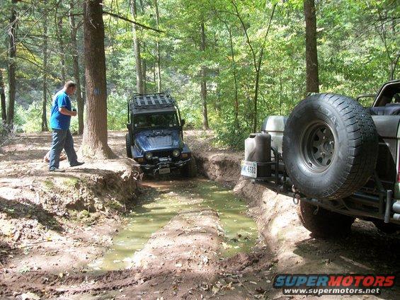 Two off-road vehicles, one blue Jeep and a rear view of another vehicle, drive through a muddy, wooded trail