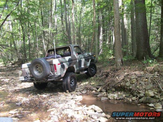 Camouflaged Ford Bronco driving through a rocky stream in a forest. Spare tire mounted on the rear