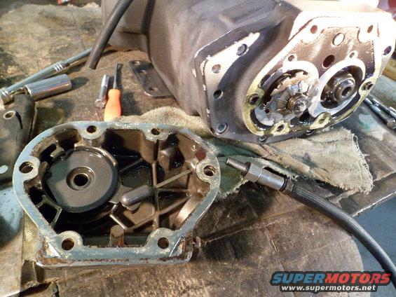 Disassembled motorcycle transmission with tools on a workbench. The transmission case is open, revealing internal gears and components