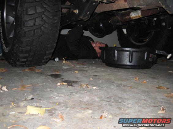 Person in black clothing lying on concrete floor under a vehicle, with a black oil drain pan nearby. Large tire visible on left