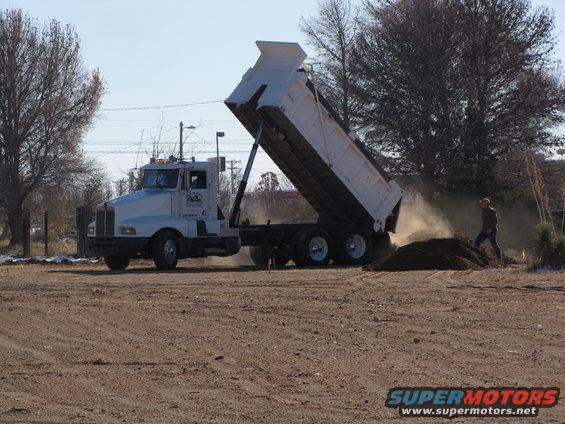 White dump truck with its bed raised, dumping dirt. A person stands nearby