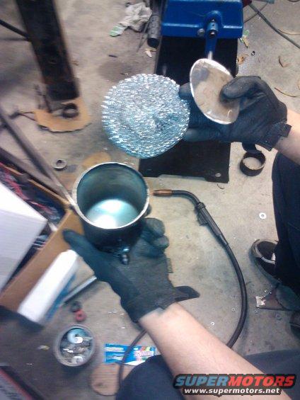 Hands in black gloves hold a metal cylinder and a metal disc with a scouring pad, with a welding torch nearby