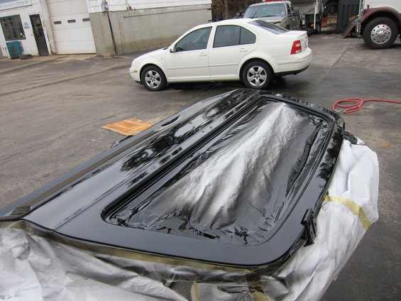 Black sunroof panel covered in plastic wrap, with a white sedan and truck in the background