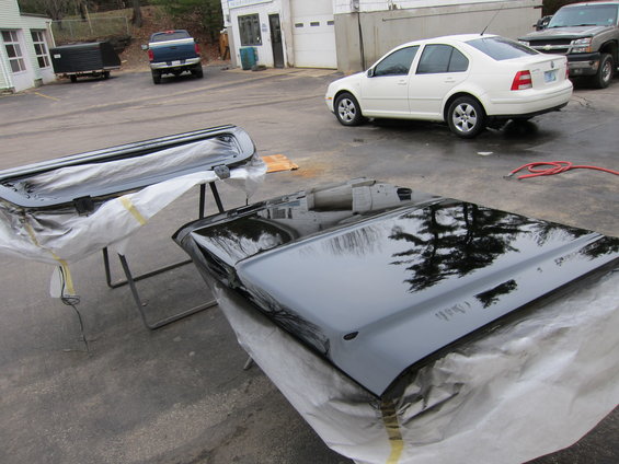Two car doors, one on a stand and one on a table, covered in plastic and being painted black. A white sedan and two trucks are in the background