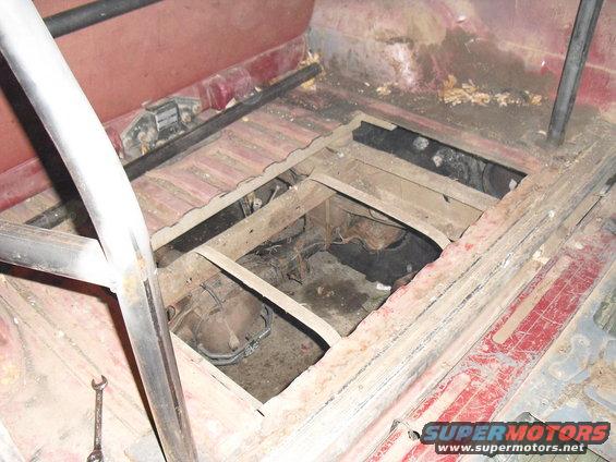 Interior floor of a vehicle with a large opening revealing the transmission and undercarriage components. Roll cage tubing is visible