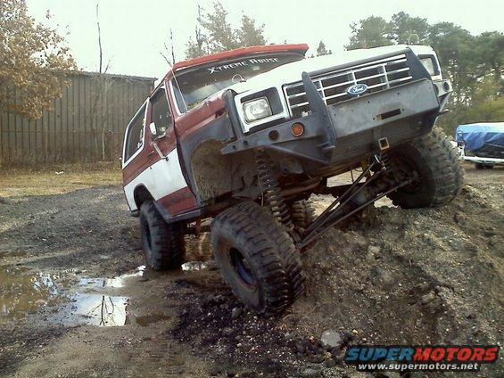 Ford Bronco with lifted suspension and large tires climbing a dirt mound. Red and white paint, custom bumper, and 'X-TREME ABUSE' sticker on windshield