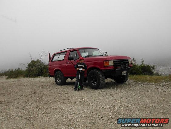 Red Ford Bronco from the late 1980s parked on a dirt road with a child and scooter