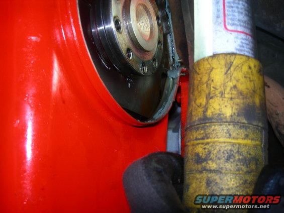 Close-up of a red automotive part with a bolted metal component and a yellow cylindrical object. Supermotors.net logo visible