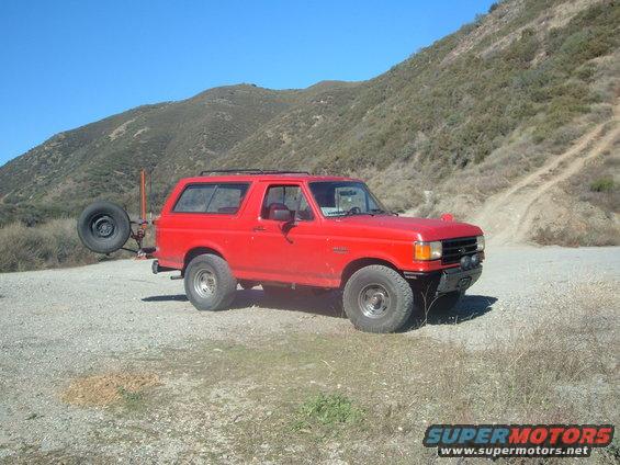 Red Ford Bronco parked on dirt road with spare tire on back. Hills in background