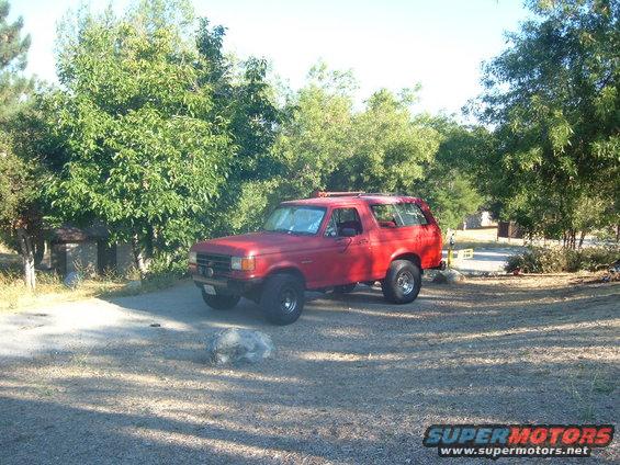 Red SUV parked on gravel, side and front visible. Trees and a small building in background