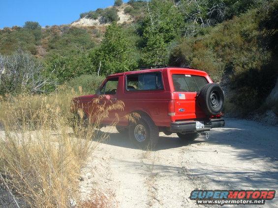 Red Ford Bronco parked on a dirt road with dry grass in the foreground and hills in the background