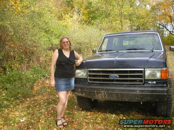 Woman in black top and denim skirt leans on a dark blue Ford Bronco. "4x4" badge visible on front