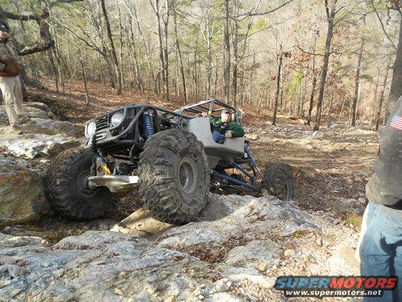 Off-road buggy with large tires navigating rocky terrain. Driver in green jacket visible