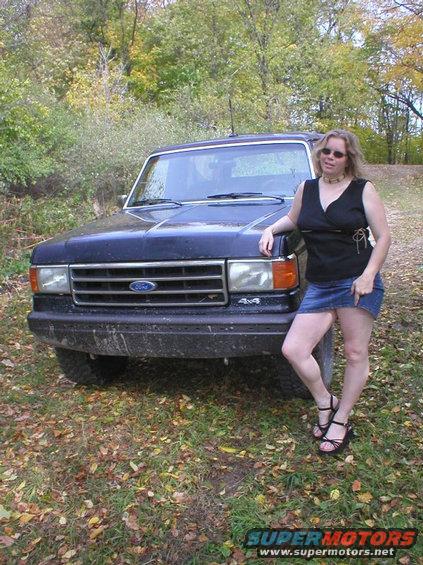 Dark SUV with a chrome grille and rectangular headlights, parked on a dirt road with fallen leaves. A woman in a denim skirt stands beside it