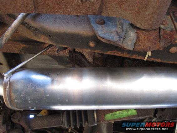 Close-up of a shiny exhaust pipe and rusty undercarriage components. Green sticker with text is visible