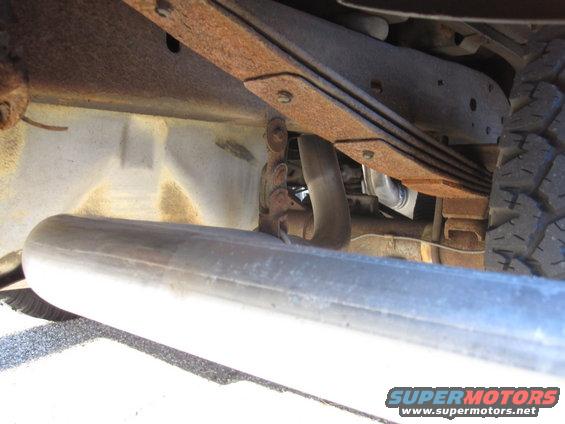 Close-up of rusty leaf springs and exhaust pipe under a vehicle. Tire tread visible on the right