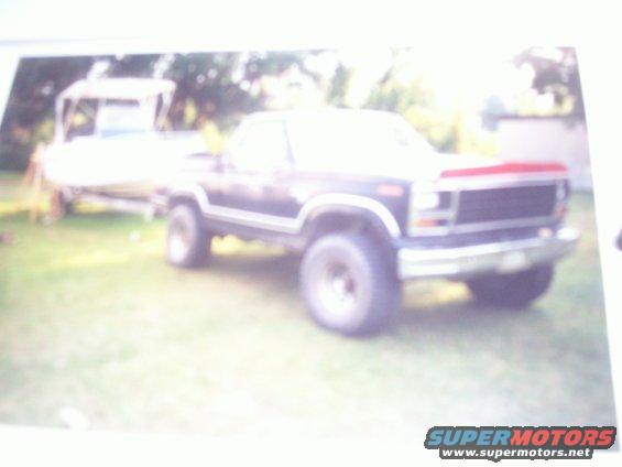 Dark blue Ford Bronco with chrome trim and large tires, parked on grass next to a boat on a trailer