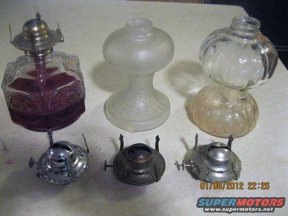 Three glass oil lamps and three metal bases on a table. One lamp is purple glass with a metal top. The other two are clear glass