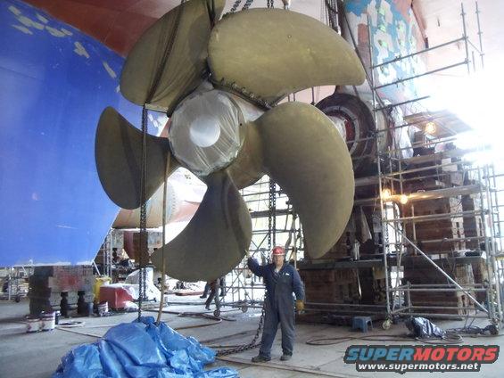 guy in jumpsuit next to huge ship propeller being worked on