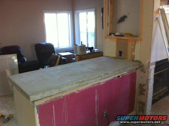 kitchen counter with concrete top, pink paneling, and exposed framing. Two chairs and windows in background