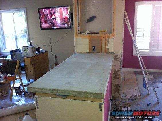 Countertop under construction with concrete surface and pink side panel, with a TV on the wall and construction materials nearby
