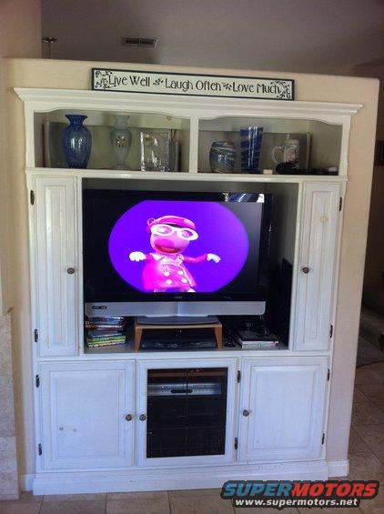 Entertainment center with TV showing cartoon character, decorative vases on shelves above, and storage cabinets below. Text on wall says 'Live Well Laugh Often Love Much'