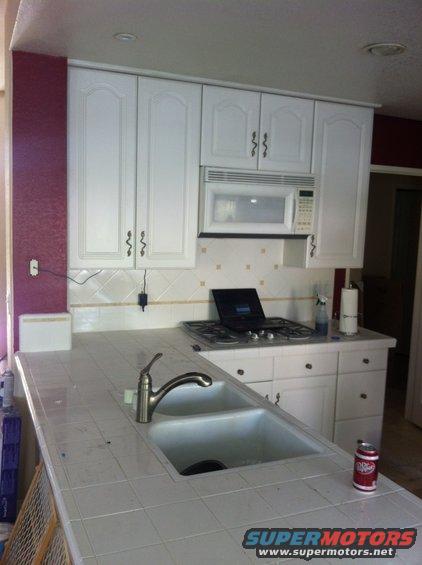 Kitchen counter with a double sink, chrome faucet, white tiled backsplash, white cabinets, a gas stove with a laptop on it, and a microwave above