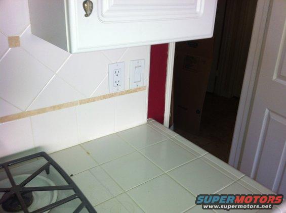 Kitchen counter with white tiled backsplash, electrical outlets, and a stove grate. A cabinet is above the counter