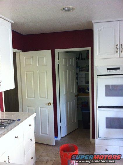 Kitchen interior with white cabinets, double ovens, and two doors, one open to a pantry