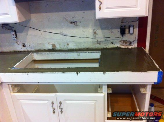Newly poured concrete countertop with sink cutout, above white kitchen cabinets. Supermotors.net logo visible