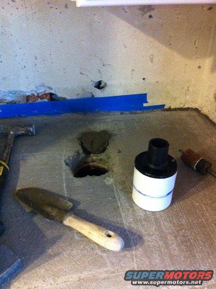 Concrete floor with a hole, a trowel, a hammer, and a white cylindrical object with a black top