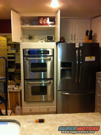 Kitchen scene with stainless steel double oven and refrigerator, open cabinet above oven, and a screwdriver on the counter