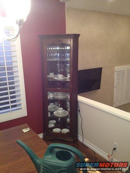 Corner curio cabinet filled with plates, bowls, and glasses. A watering can sits on a wooden table in the foreground