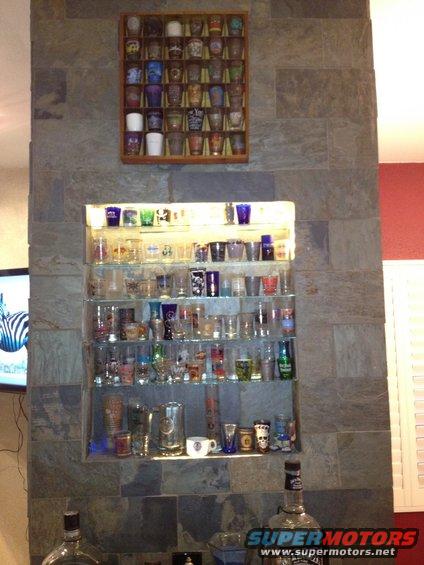 Wall display with shelves filled with shot glasses and barware, some bottles on the counter below