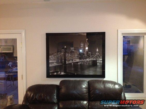 Framed picture of Brooklyn Bridge and Manhattan skyline hanging above a leather couch. Two dogs are visible through a glass door on the left