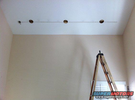 Wooden A-frame ladder positioned in front of a window with blinds, near a ceiling with four circular openings
