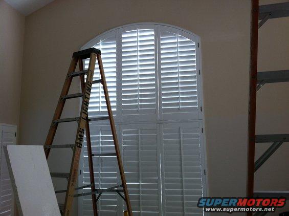 wooden ladder leaning against large arched window with white plantation shutters