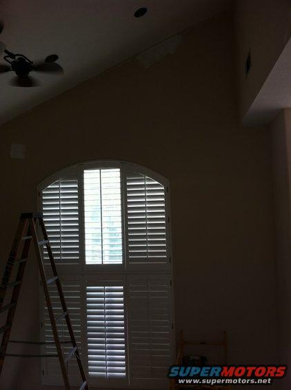 Interior scene with a tall arched window covered by white shutters, a wooden ladder leaning against the left side, and a ceiling fan