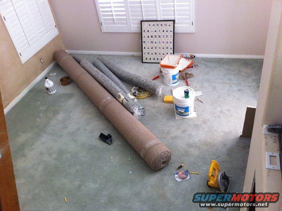 Room with rolled carpets, paint buckets, rollers, and a circular saw on a concrete floor