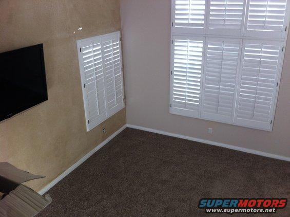 Interior room with brown carpet, tan textured walls, a flat-screen TV, and white plantation shutters on two windows