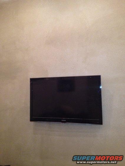 Black flat-screen television mounted on a textured beige wall, with a Supermotors.net logo in the lower right corner