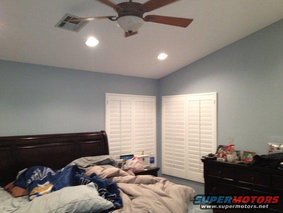 Bedroom with unmade bed, dark wood headboard, blue walls, white plantation shutters, ceiling fan, and dresser